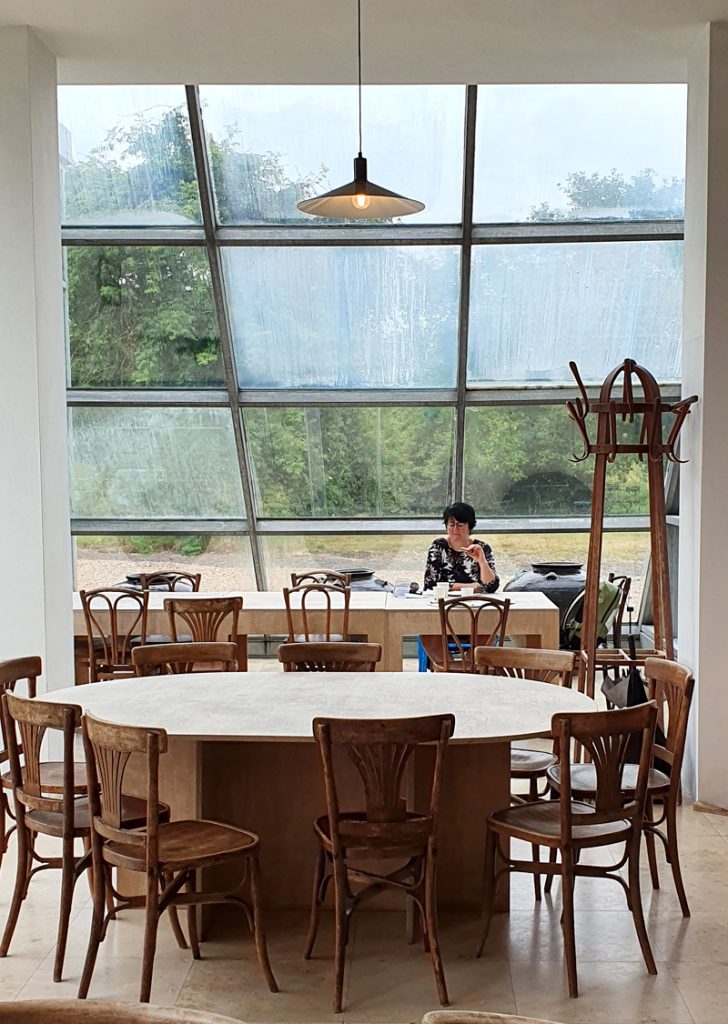 Eine Frau sitzt vor einer verglasten Wand in einer Cafeterie. Im Vordergrund Tische und Stühle.